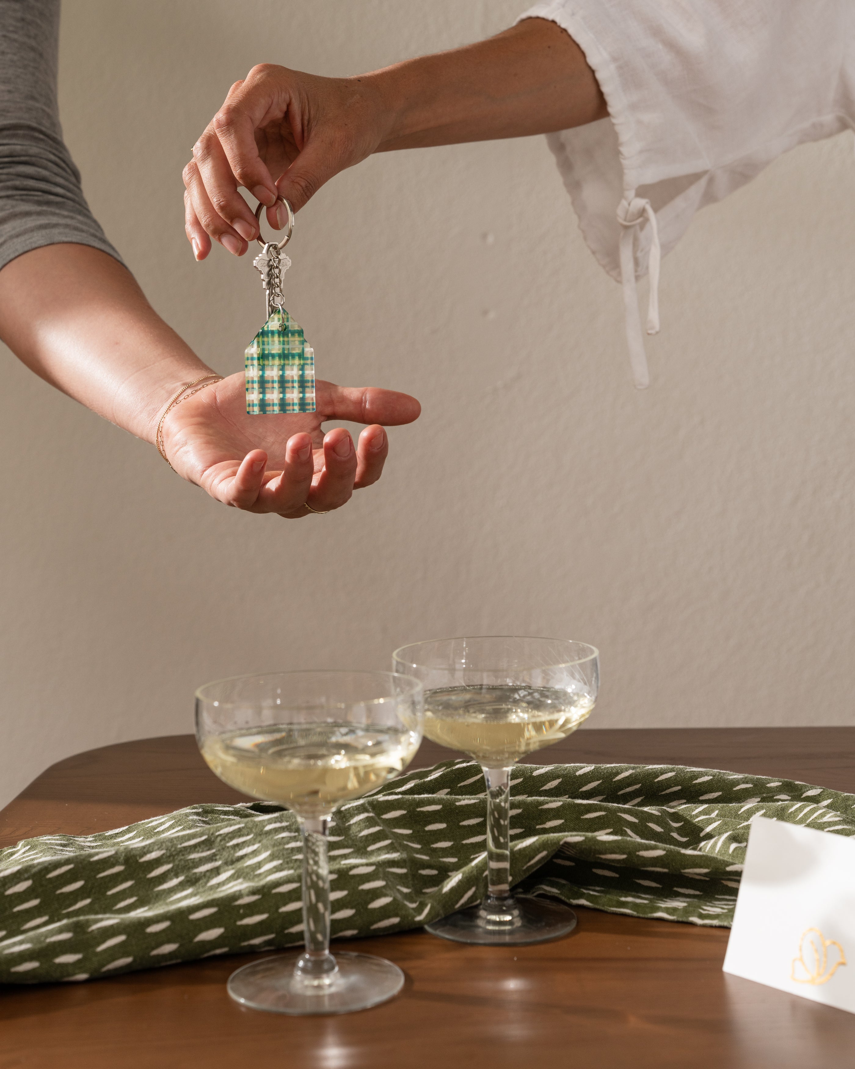 Person holding a decorative keychain above two glasses of white wine on a table with a green patterned cloth.